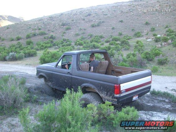 Dark Ford Bronco driving through mud and brush on a dirt road with a hill in the background