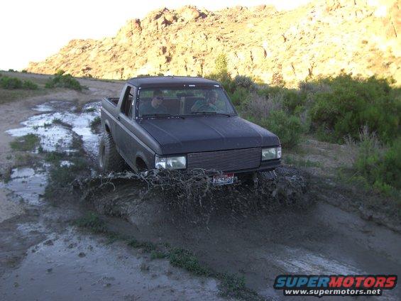 Black 1987 Ford Bronco splashing through a muddy trail with two people inside