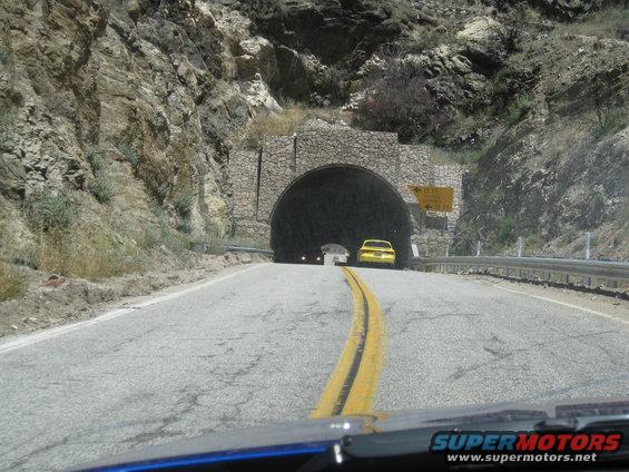 Yellow sedan seen from behind, approaching a stone tunnel entrance on a mountain road