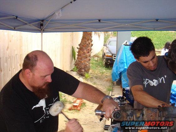 Two men working on a vehicle axle assembly outdoors under a canopy. A dark SUV is visible in the background