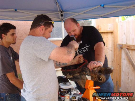 Axle assembly on orange jack stand, two men working on it, third man watches