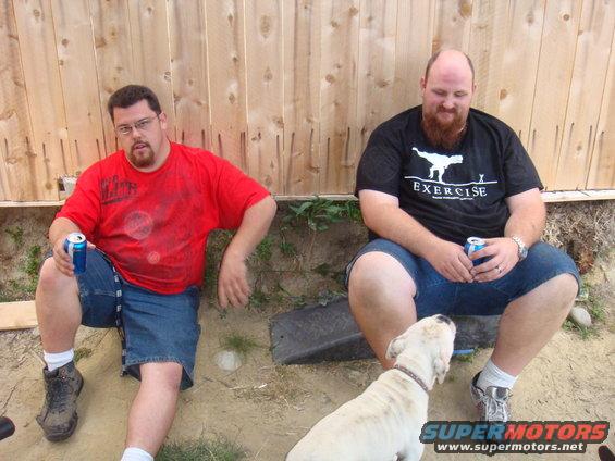 Two men sit by a wooden fence, one holding a can, with a white dog looking up at the other man. "SUPERMOTORS" logo visible