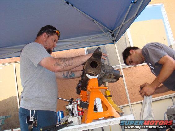 Two men working on a vehicle axle assembly supported by orange jack stands