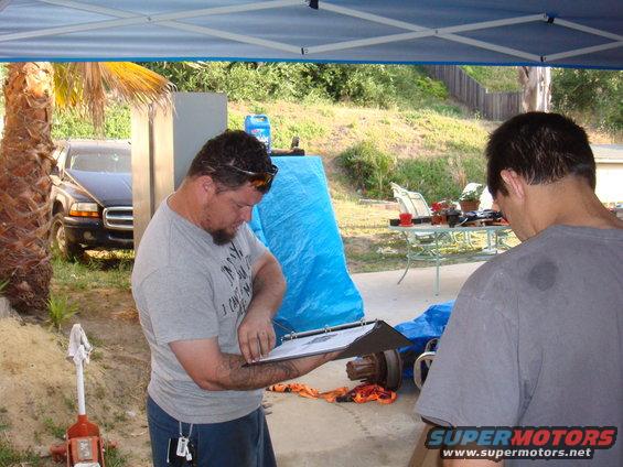 Two men stand under a canopy, one holding a clipboard, with a vehicle axle visible on the ground