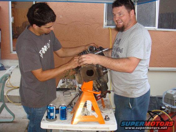 Engine part on orange jack stands with two men working on it, two cans of soda on table