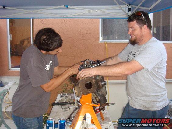 Two men work on a vehicle axle assembly on an orange jack stand