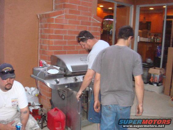 Three men stand around a stainless steel gas grill on a patio with a brick structure
