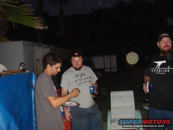 Three men stand around outside at night, holding drinks. One man wears a shirt that says 'I'm sorry, I can't hear you over the sound of how awesome I am.'