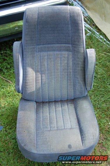 Fabric car seat with armrests, sitting on grass. Part of a vehicle bumper is visible behind it