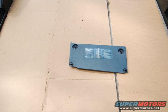 Dark gray rectangular vehicle part with white text and diagrams, lying on cardboard