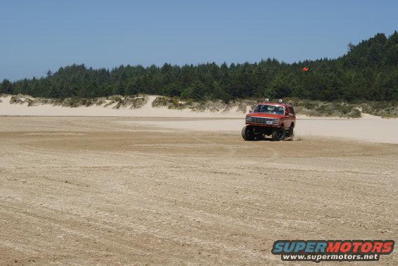 Red SUV with off-road tires driving on a sandy beach with trees and dunes in the background