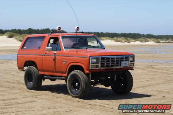 Orange Ford Bronco with roof rack and off-road tires driving on a sandy beach