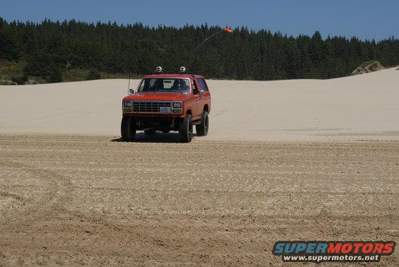Orange 1983 Ford Bronco driving on sand dunes with an orange flag on an antenna