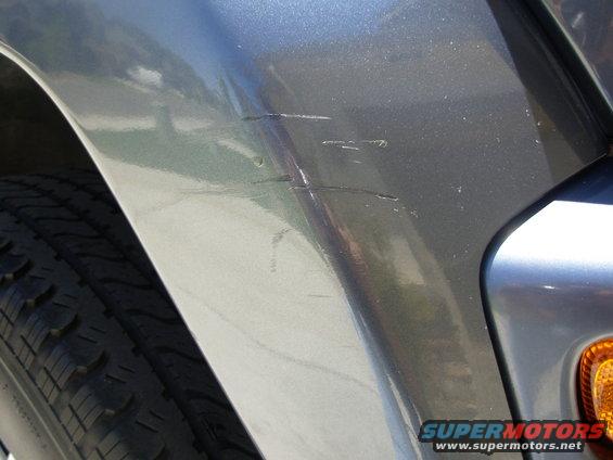 Gray vehicle body panel with visible scratches near the wheel well and a portion of the tire. Text 'SUPERMOTORS' is visible at the bottom right