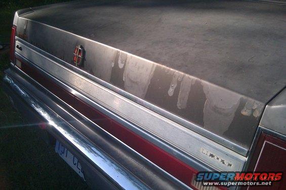 Rear of a Lincoln Town Car, silver trunk lid with water stains, chrome trim, red taillight strip, license plate visible