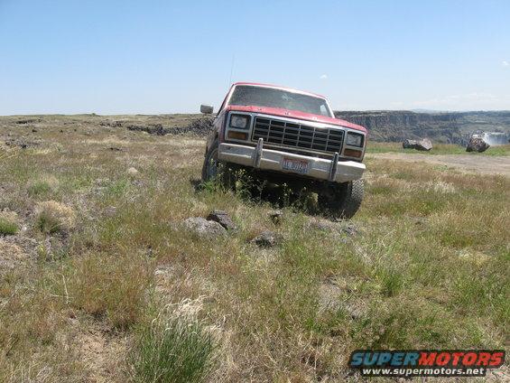 Red pickup truck with chrome grille and bumper on grassy, rocky ground. License plate visible