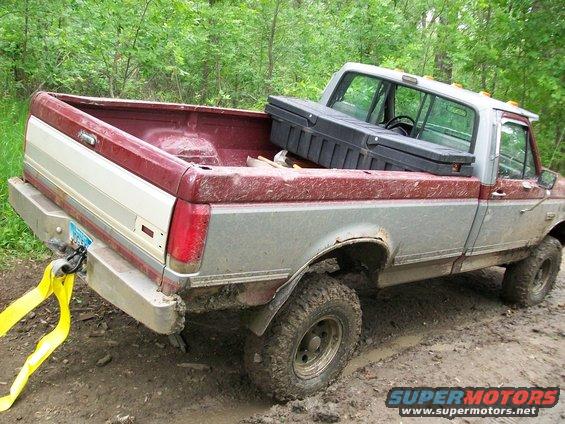 Rear view of a red and silver Ford pickup truck stuck in mud, with a yellow tow strap attached to the bumper