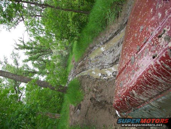 Red truck's fender and door covered in mud, driving on a muddy track in a wooded area