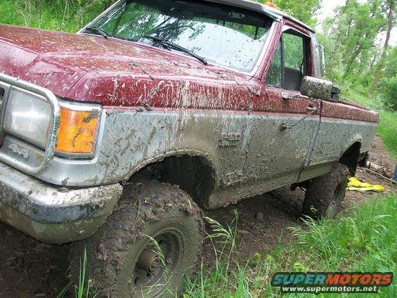 Muddy red and silver pickup truck stuck in dirt with tall grass around