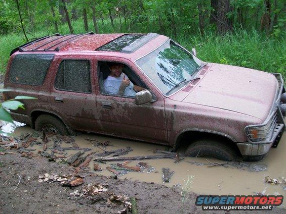 Red SUV stuck in mud, driver giving thumbs up. Visible sunroof and aftermarket bumper