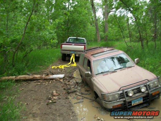 Tan SUV stuck in mud, being pulled by a pickup truck with a yellow strap on a wooded trail