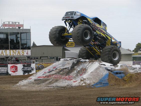 Monster truck 'Obsessed' mid-air over a dirt ramp, with other crushed cars visible below