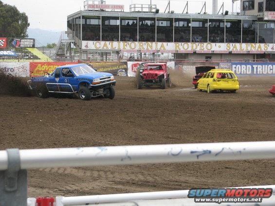 Blue pickup truck kicking up dirt on a race track, with other vehicles visible