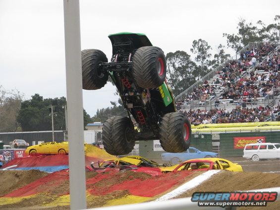 Monster truck airborne over crushed cars at an event with spectators in bleachers