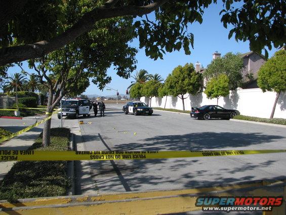 Black sports car with a spoiler parked on a residential street with police tape and vehicles