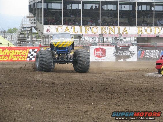 Monster truck with yellow and blue body in dirt arena, grandstands in background