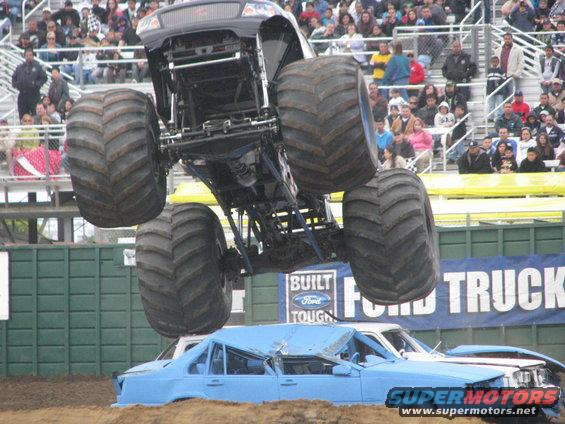 Monster truck airborne over crushed cars at an event. Crowd in background