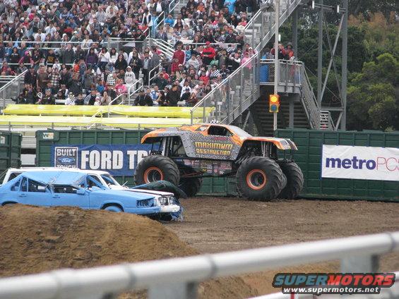 Crushed blue sedan and orange monster truck with 'Maximum Destruction' text, in a dirt arena with spectators