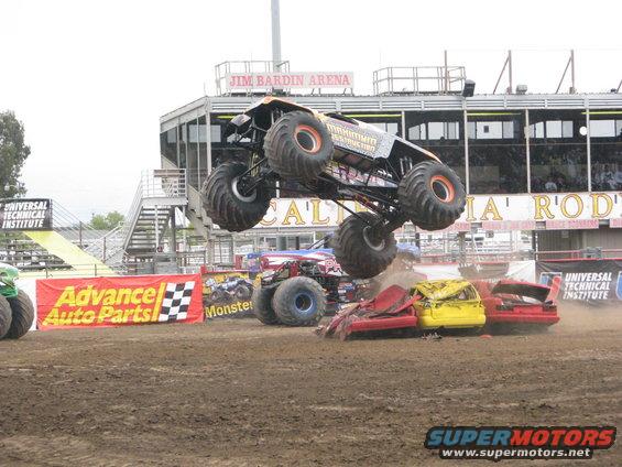 Monster truck airborne over crushed cars at a stadium event. Other monster trucks and a banner for Advance Auto Parts are visible