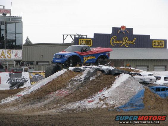 Blue monster truck with Superman cape on dirt ramp, other cars in background
