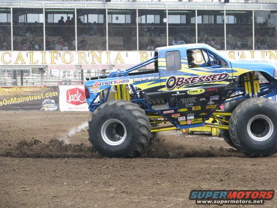 Monster truck named Obsessed driving on dirt track, kicking up dust. Big tires, blue and yellow paint scheme