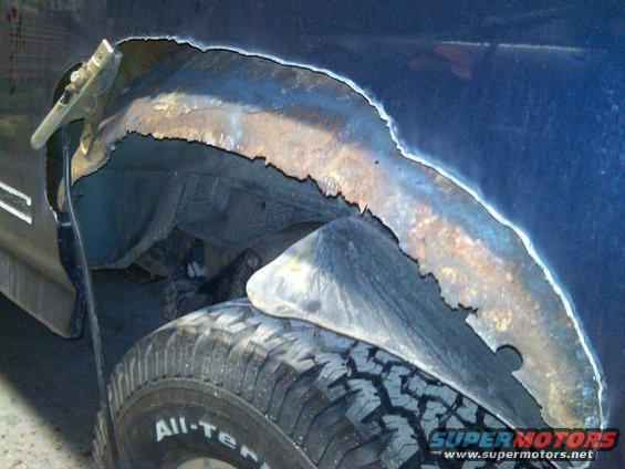 Close-up of a rusted-out wheel well on a dark blue vehicle, with a tire visible below