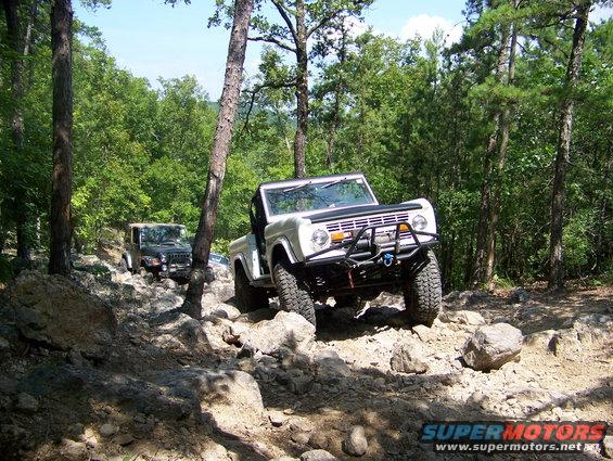 White Ford Bronco and dark Jeep navigating a rocky, wooded trail