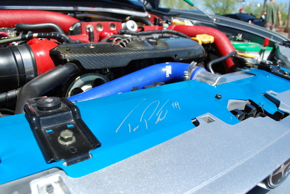 Engine bay with blue and red hoses, carbon fiber cover, and a signature on a blue plate