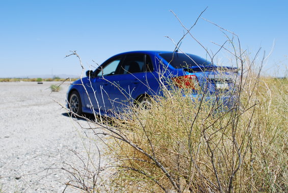 Blue sedan parked on a gravel lot with dry brush in the foreground
