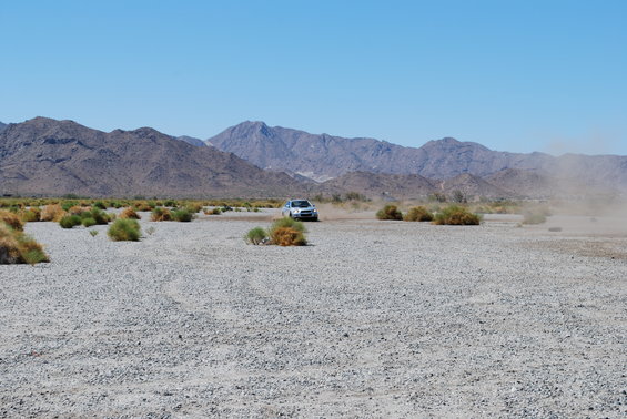 Silver sedan driving on a gravelly plain with mountains in the background