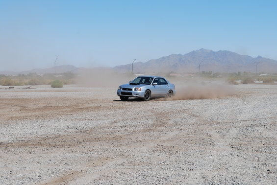 Silver 2005 Subaru Impreza sedan drifting on a dirt lot, kicking up dust. Mountains in background