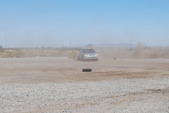 2005 Subaru Impreza driving on a dirt road, kicking up dust. A tire lies on the ground in the foreground