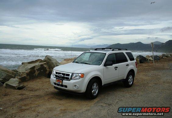 White Ford Escape SUV parked on a dirt road near the ocean, with a rocky shoreline and mountains in the background