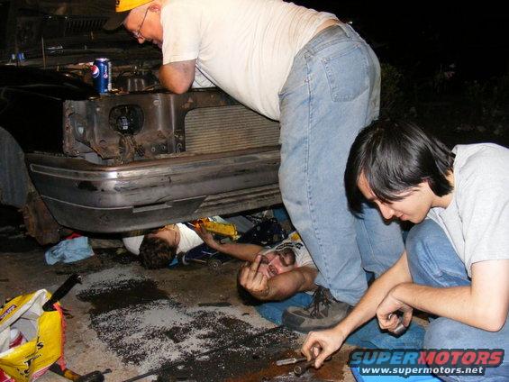 Guys working on a car's front end, one is under it giving the finger