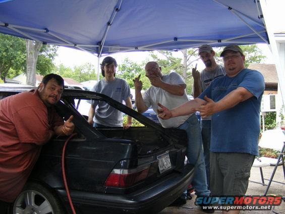 Five men gathered around the open trunk of a dark-colored 1989 Ford Escort hatchback