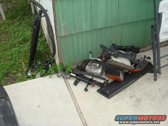 Pile of car parts including an AC compressor, headlights, and bumper trim next to a green wooden wall