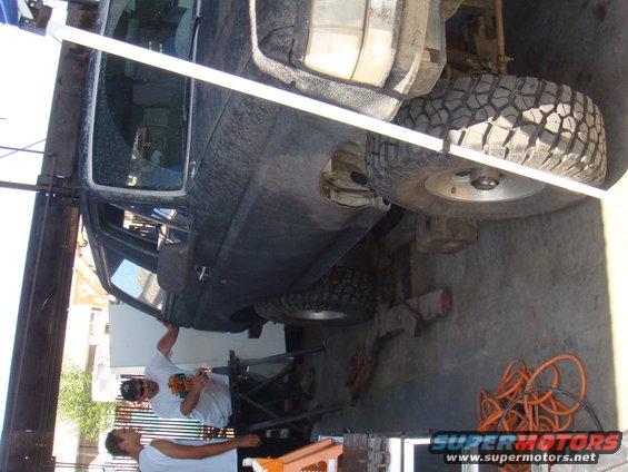 Side view of dark SUV on lift with men working underneath. No year visible