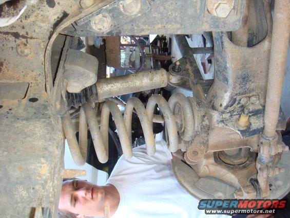 Underside view of a vehicle's suspension system, focusing on the coil spring and shock absorber assembly. A person is visible looking at the components