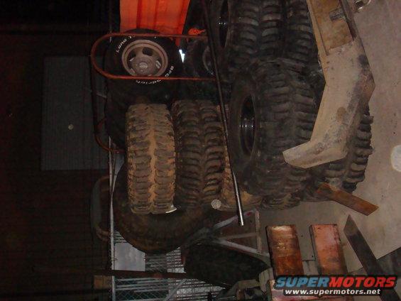 Stack of large off-road tires, some with mud on them. One tire has a silver rim with text