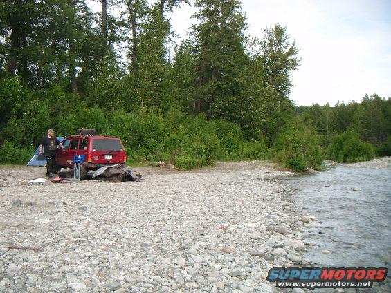 Red 1998 Jeep Cherokee parked on a rocky riverbank with a person and tent nearby